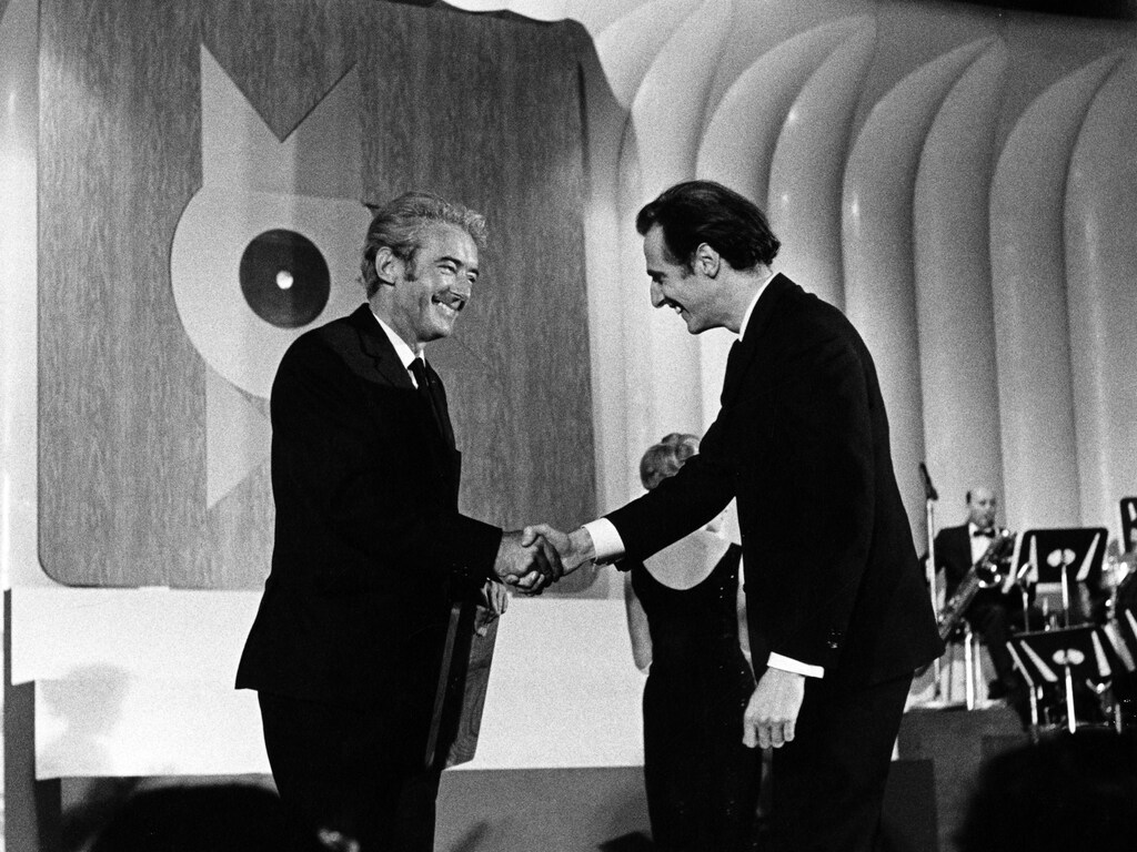 Sur une scène, l'auteur-compositeur québécois Félix Leclerc serre la main de l'auteur-compositeur québécois Gilles Vigneault en 1965.