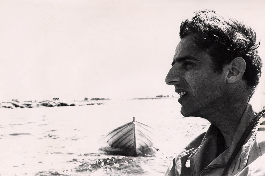 Gilles Vigneault regarde au loin. Il est devant l'eau, on voit une barque à l'arrière.